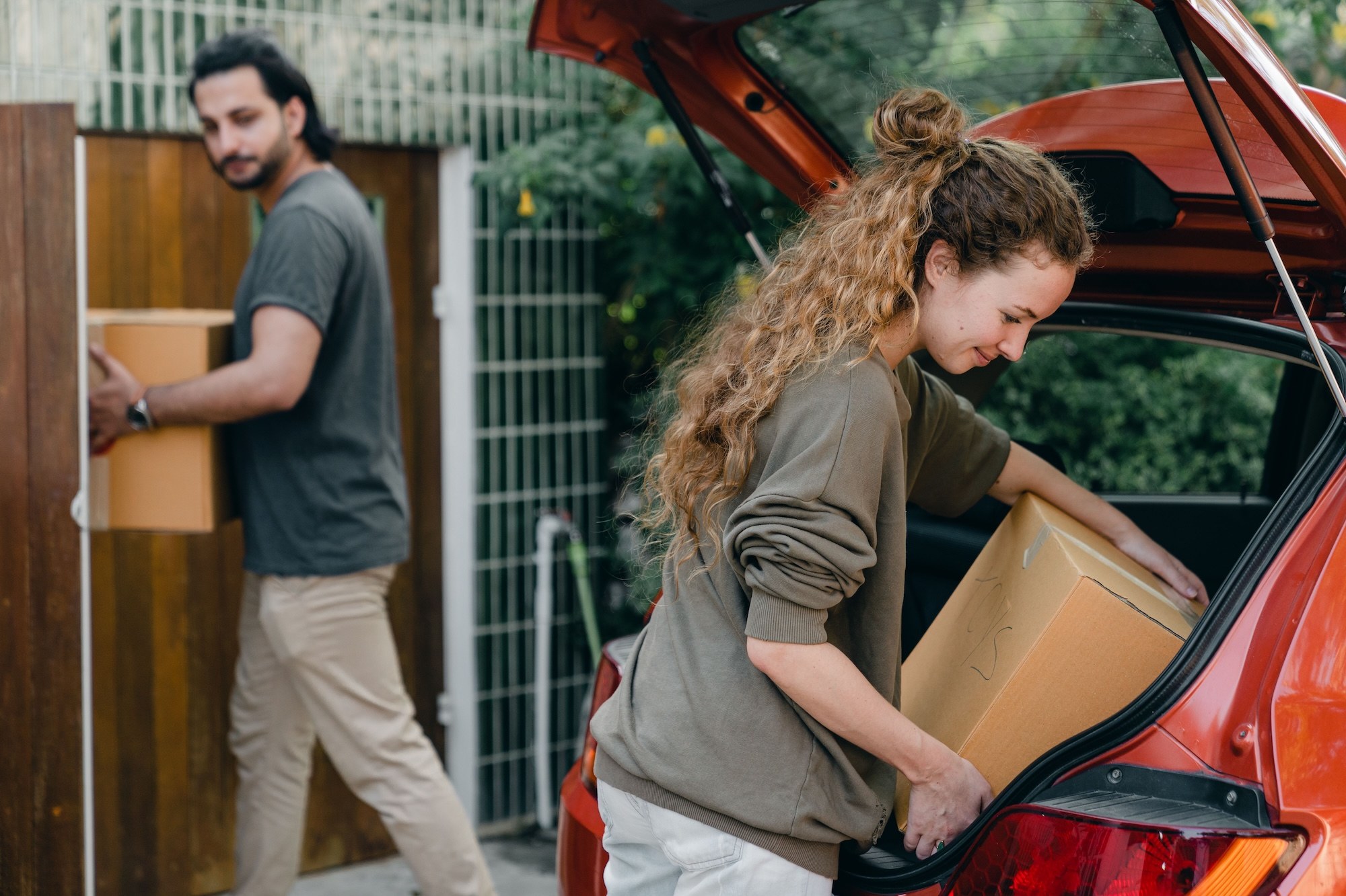 A couple carrying cardboard moving boxes from the open hatch of a red car through a wooden gate.